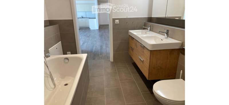 Modern bathroom with a bathtub, double sink, and toilet. The space features wooden cabinets, tiled walls, and a view into an adjacent kitchen area.
