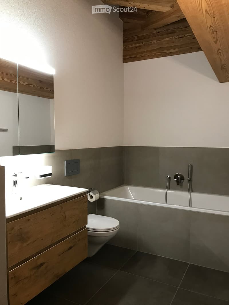 Modern bathroom with a wooden vanity, white sink, toilet, bathtub, and built-in lighting. Dark tiles on the floor and walls.