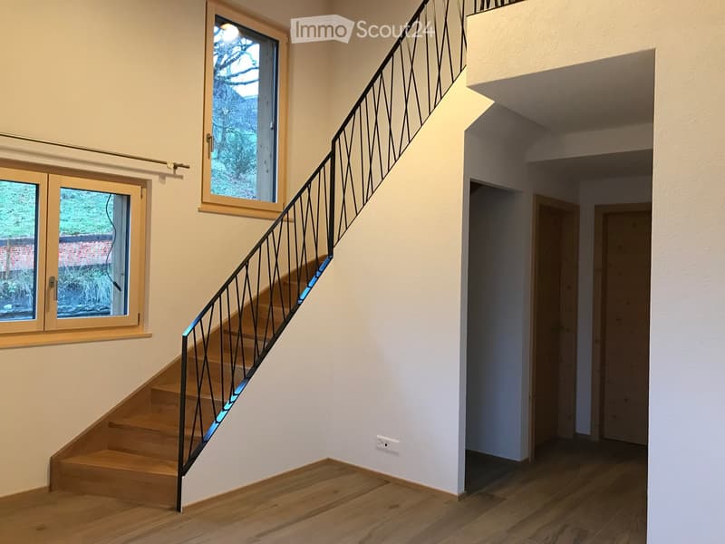 Staircase with wooden steps and metal railing, leading to a hallway with wooden flooring and white walls. Windows on the left side provide natural light.