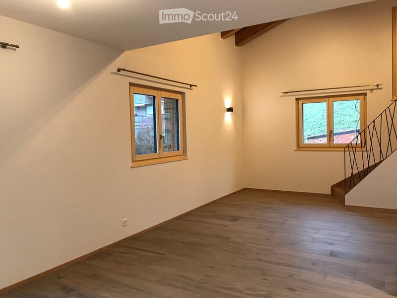 Empty room with wooden flooring, white walls, and two windows with wooden frames. A staircase with a metal railing is visible on the right side.