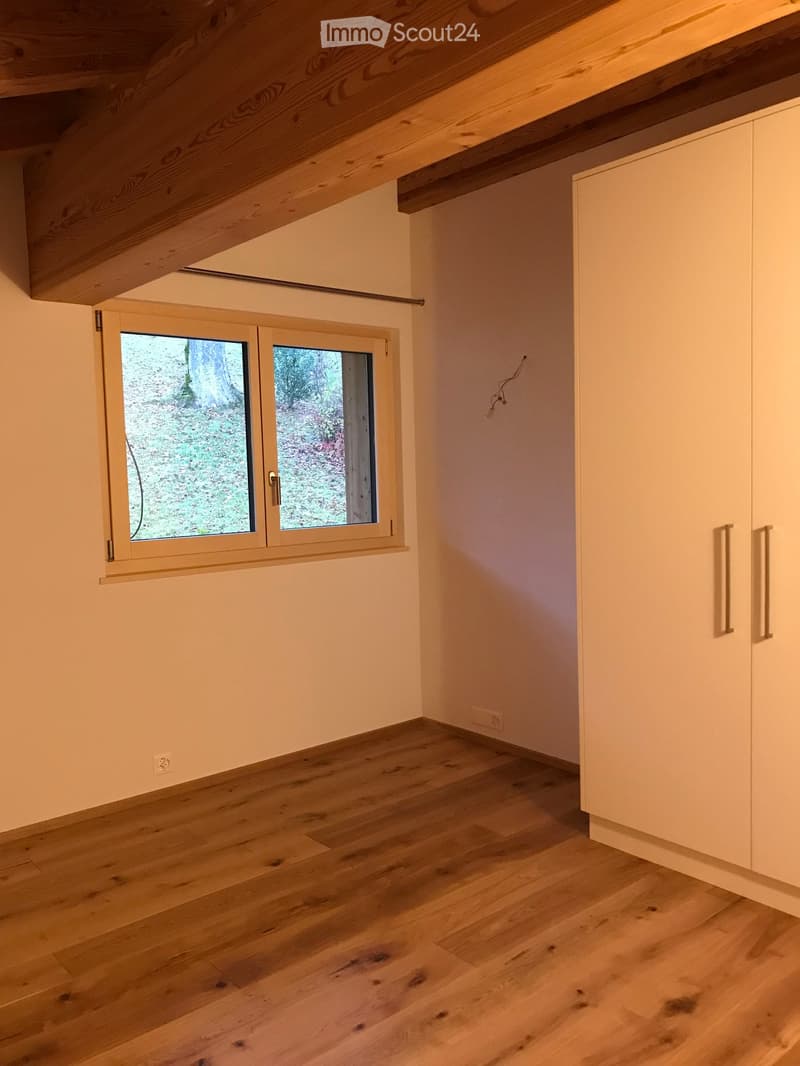 Empty room with light wooden flooring, white walls, built-in white cabinets, and a large window with a view of greenery outside.