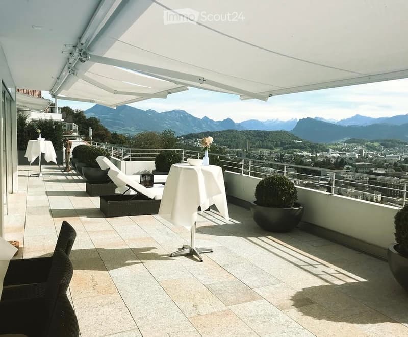 A spacious balcony with a retractable awning, offering a scenic mountain view. It features a round table with a white tablecloth, a lounge chair, and several potted plants.