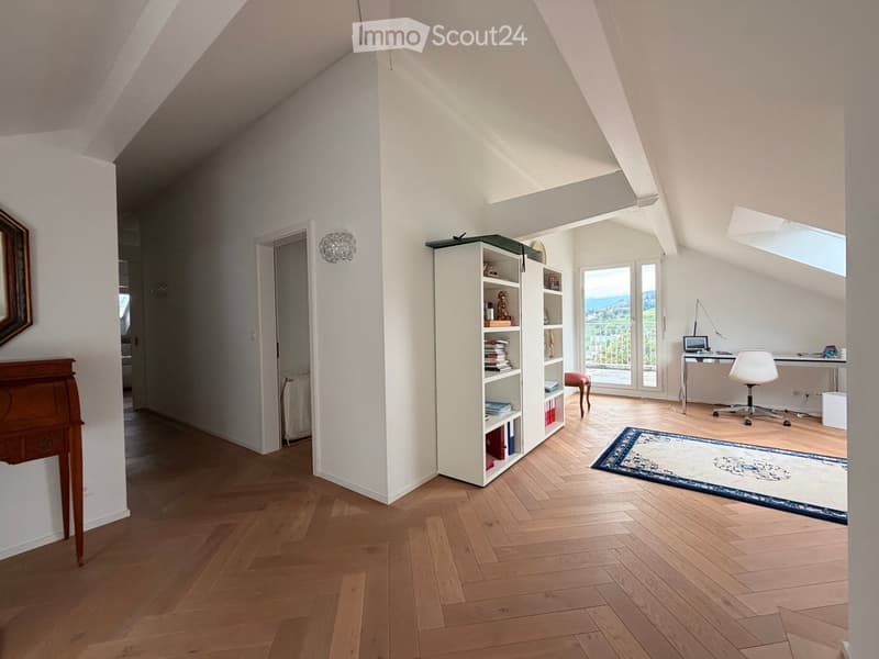 Bright attic room with wooden flooring, large windows, and a view. Features a desk with a chair, a bookshelf, a small table, and a mirror on the wall.