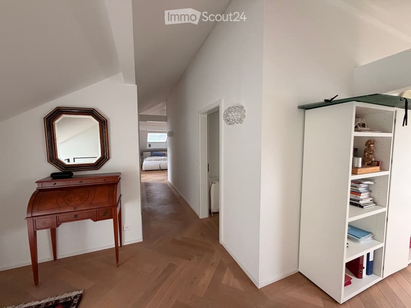 A narrow hallway with wooden flooring, white walls, and a slanted ceiling. Features include a wooden desk with a mirror, a white shelving unit with books and decorative items, and a view into an adjacent bedroom with a bed and window.