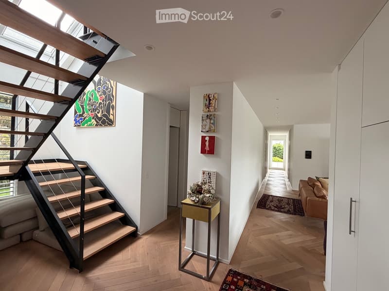 Modern hallway with wooden flooring, a staircase leading upwards, and artwork on the walls. A small table with a floral arrangement is placed near the staircase, and a living area with a sofa is visible at the end of the hallway.
