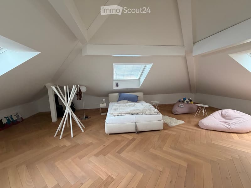 Modern attic bedroom with wooden flooring, slanted ceilings, and skylights. Features a white bed with blue pillows, a beige bean bag chair, a white floor lamp, and a white coat rack.