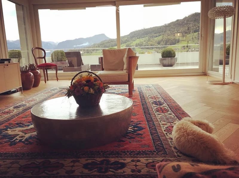 Bright living room with large windows offering a scenic mountain view, featuring a sofa, coffee table, armchair, floor lamp, decorative vases, and a flower arrangement on the coffee table.