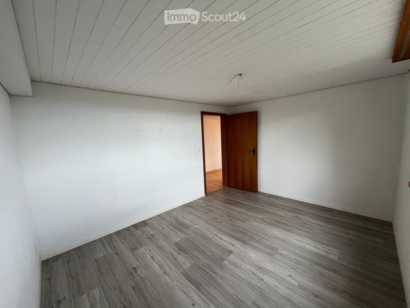 Empty room with white walls, grey wood flooring, and a wooden door leading to another room.