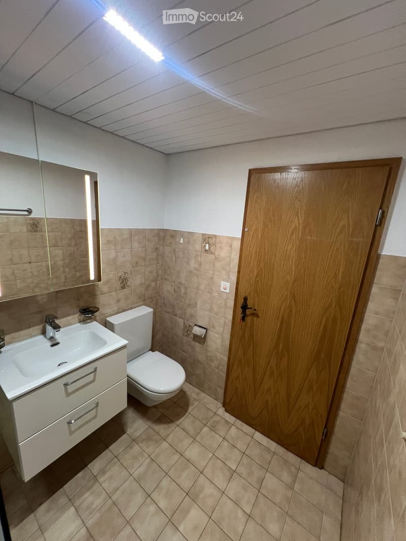 Modern bathroom with beige tiles, white sink, toilet, and a large mirror with built-in lighting. Wooden door visible on the right.