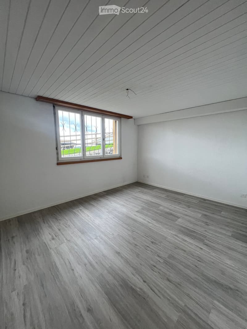 Empty room with wooden flooring, white walls, and a large window with wooden frame and metal bars.