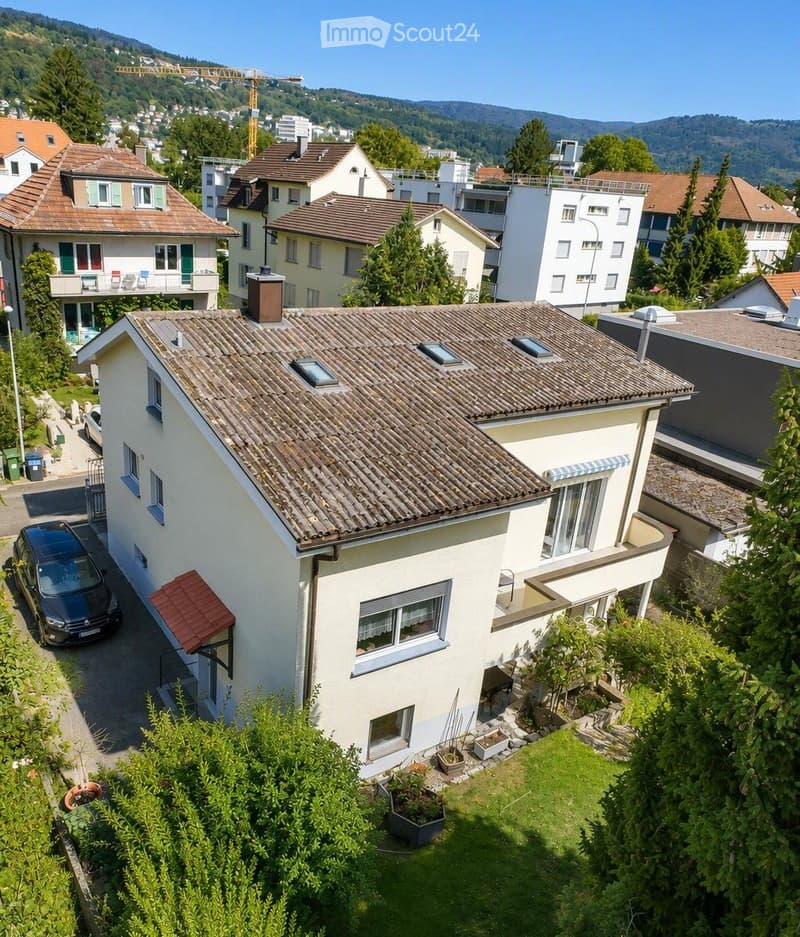 A two-story house with a tiled roof, white exterior, multiple windows, and a small garden area with potted plants.