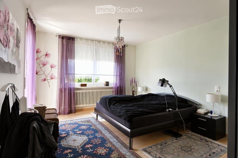 Modern bedroom with a large bed, nightstand, and a variety of lamps. The room features wooden flooring, a colorful rug, and large windows with purple curtains.