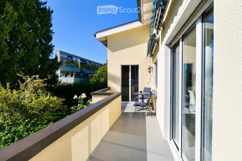 Balcony with a small table and chairs, overlooking a garden area with trees and shrubs.
