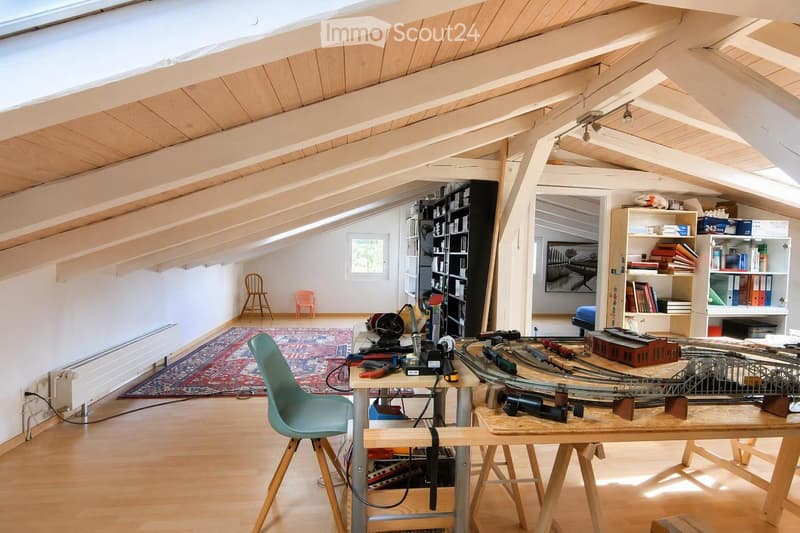 A spacious, well-lit attic room with wooden floors and exposed wooden beams. The room features a large table with a model train set, tools, and various equipment. There are shelves filled with books and storage boxes, a colorful rug, and several chairs, i