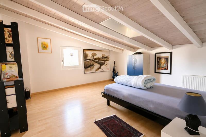 Bright bedroom with wooden floors, slanted wooden ceiling, large window, bed with blue bedding, nightstand with lamp, bookshelf, and artwork on walls.