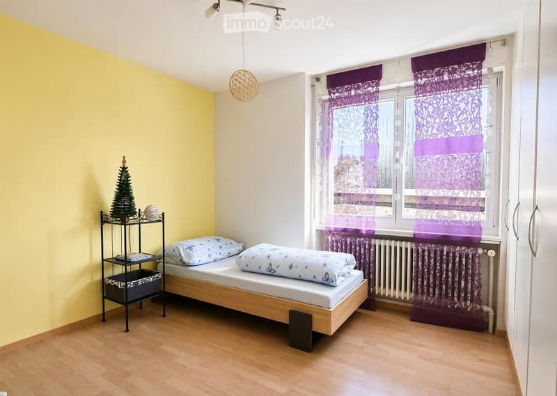 A small, cozy bedroom with a single bed, nightstand, purple curtains, and a small decorative tree on a shelf.