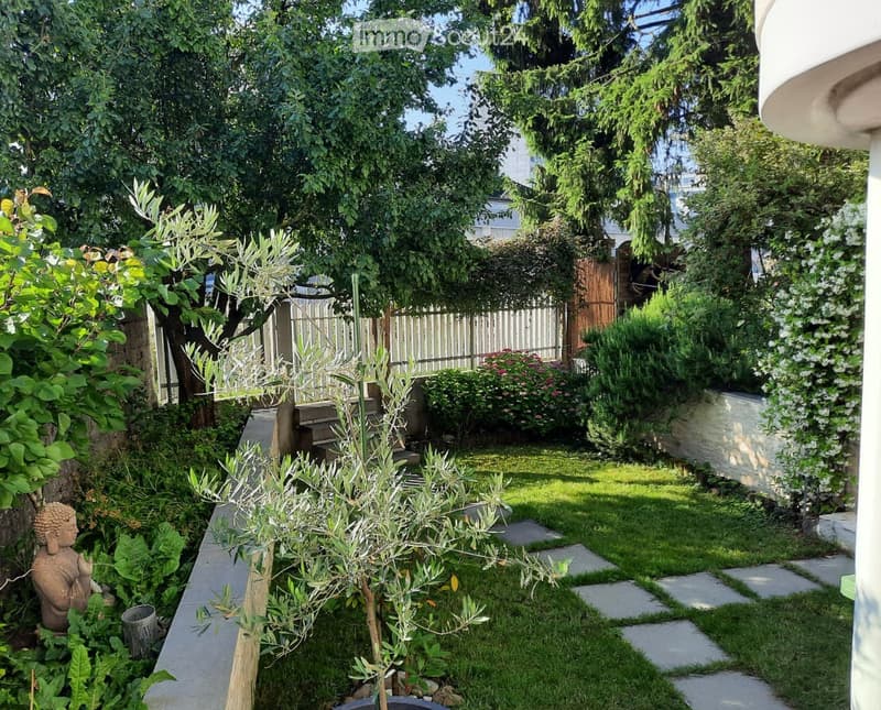 A well-maintained garden with a variety of plants, including olive trees, shrubs, and flowers. The garden features a stone pathway, a small staircase, and a wooden fence. There is also a small statue and a trash can visible.