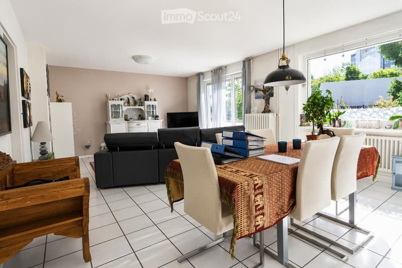 Bright living and dining area with a black sectional sofa, a dining table with chairs, a hanging pendant light, and a cabinet with decorative items. The space features large windows with sheer curtains and a view of greenery outside.