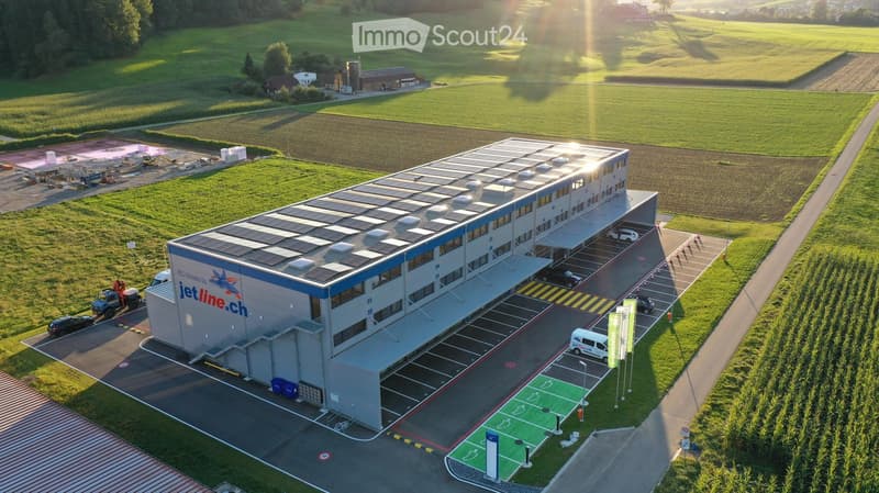 A modern industrial building with a flat roof, solar panels, and a parking lot, surrounded by green fields and agricultural structures.