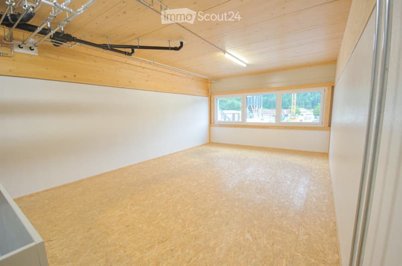Empty room with wooden flooring and exposed wooden ceiling beams, featuring large windows with a view of greenery outside.