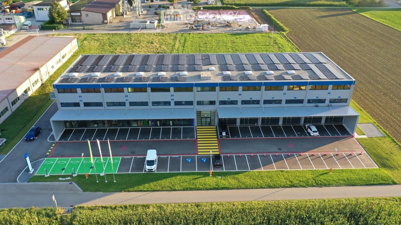 A modern industrial building with solar panels on the roof, multiple parking spaces, and a green area in front.