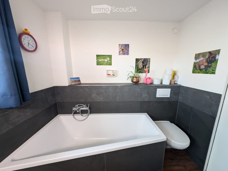 Modern bathroom with a white bathtub, toilet, dark grey tiles, and decorative items on the counter including a plant and a clock.