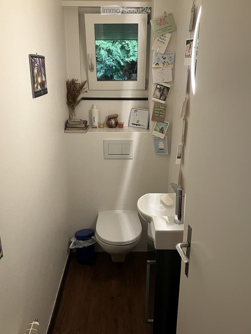 Compact bathroom with a toilet, sink, and small shelf holding various items. The room features a window with a view of greenery, and the walls are adorned with several small decorations and photos.