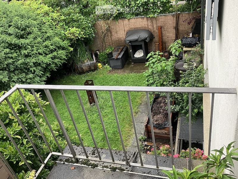 Balcony overlooking a small, well-maintained garden with a brick wall, barbecue, and various plants.