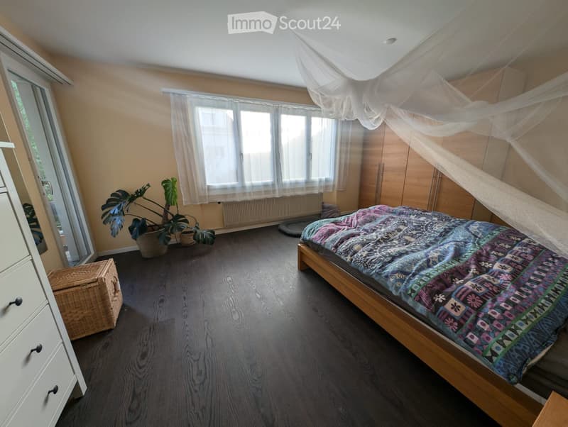 Bedroom with wooden flooring, large window with white curtains, bed with colorful quilt, wardrobe, wicker basket, and potted plants.