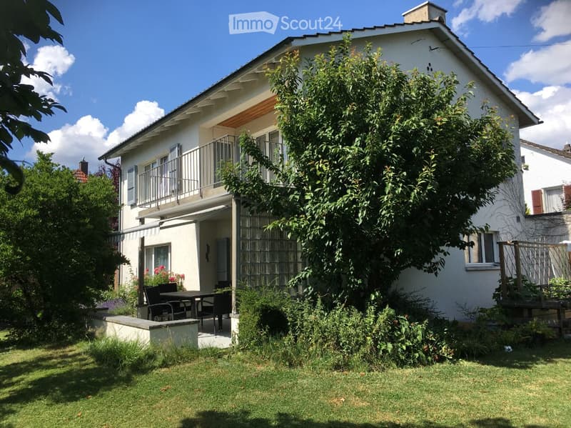 Two-story white house with a balcony, surrounded by a well-maintained garden and lawn.