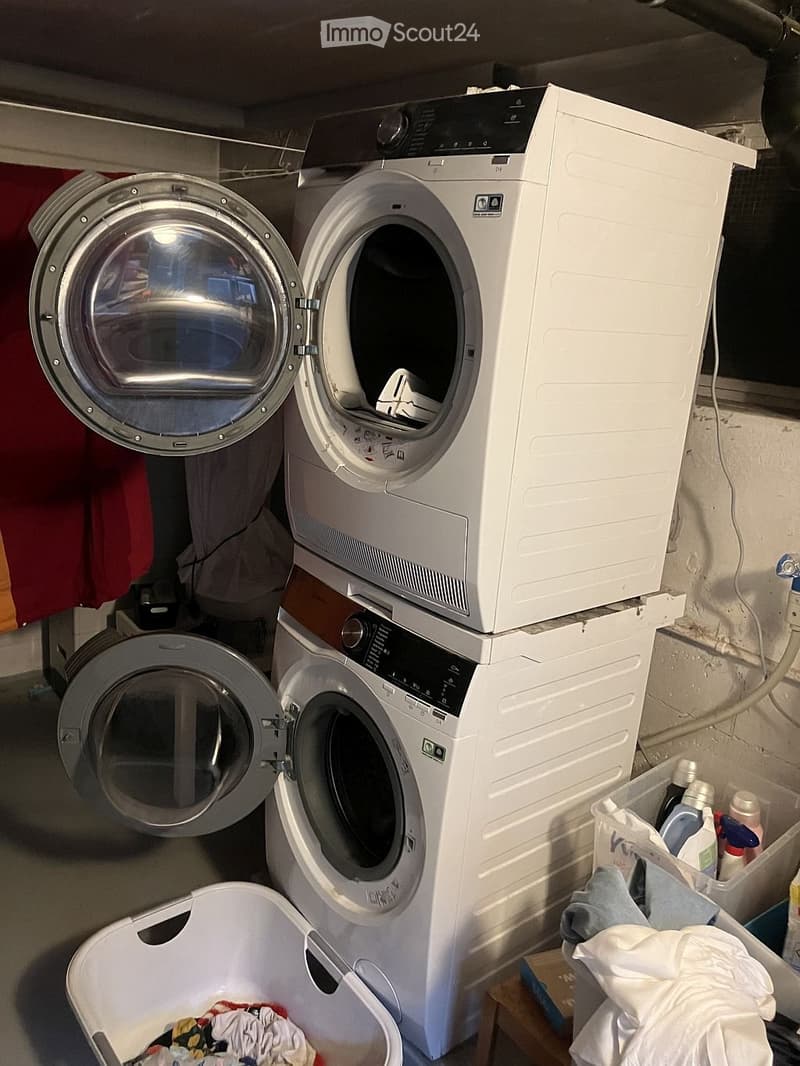 Laundry room with a stacked washing machine and dryer, both with open doors, and a laundry basket filled with clothes.
