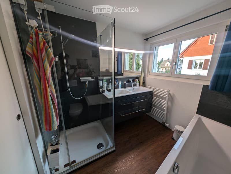 Modern bathroom with a glass-enclosed shower, double sink, mirror, radiator, and a towel hanging on the wall. The room has wooden flooring and large windows offering a view of the outside.