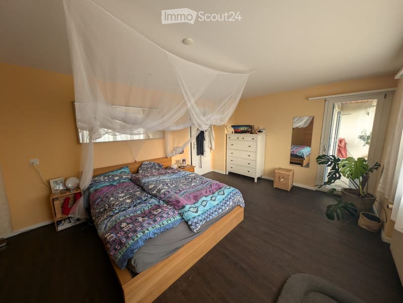 Bright bedroom with wooden flooring, double bed with colorful bedding, nightstand, dresser, mirror, and a large plant near the sliding glass door.