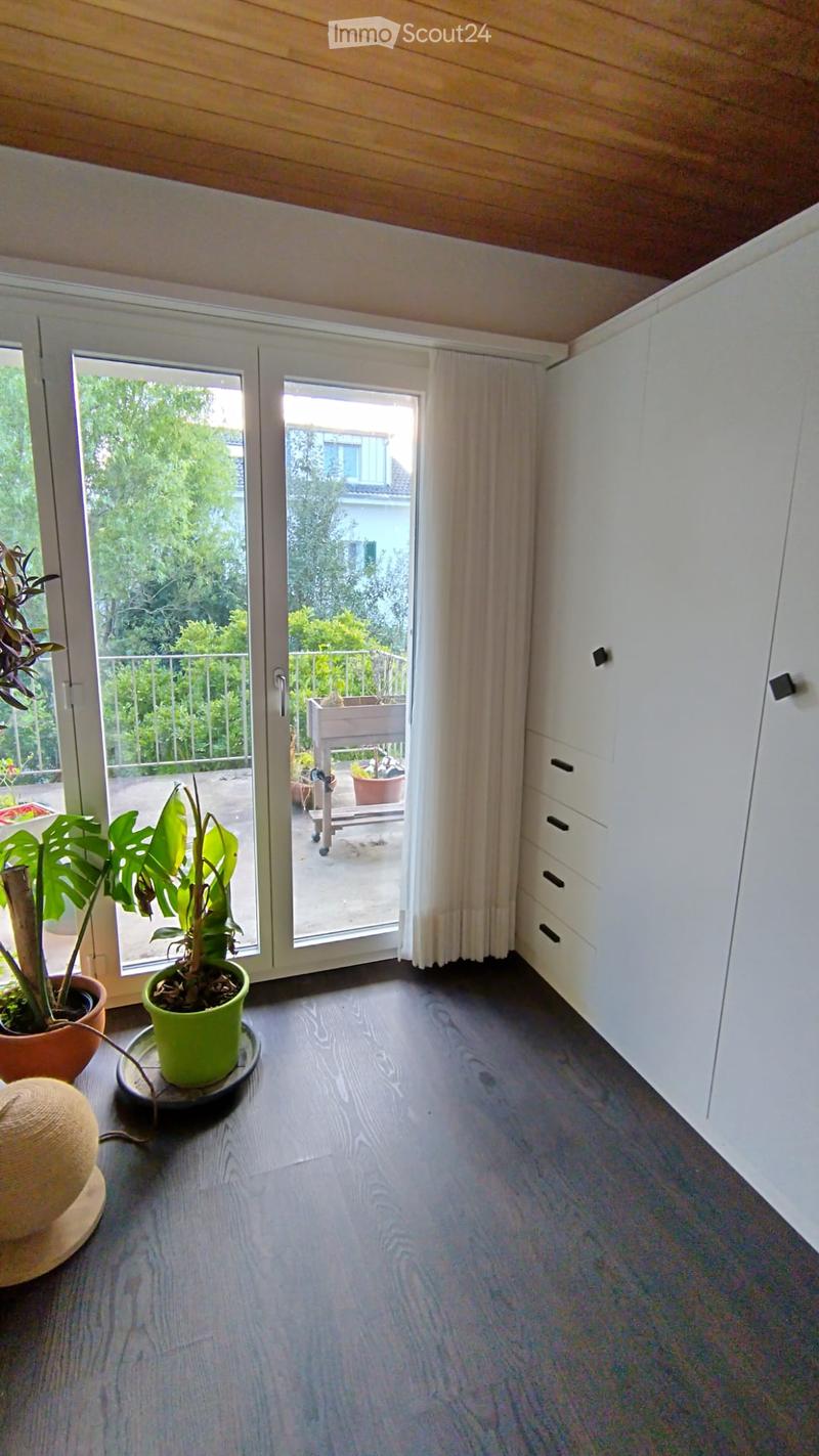 Bright room with wooden flooring, white built-in wardrobe, large sliding glass doors leading to a balcony, and potted plants.