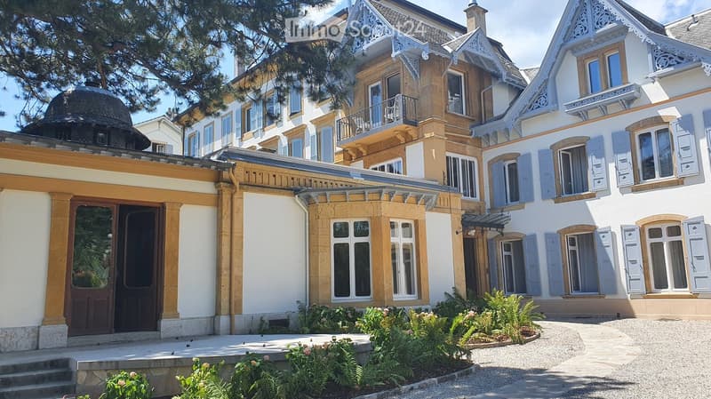 A large, multi-story residential building with ornate architectural details, including decorative gables, shutters, and a balcony. The building features a mix of light-colored walls and wooden accents, with a well-maintained garden in the foreground.