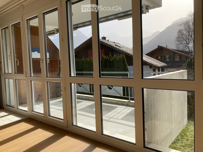 Bright, unfurnished room with large windows offering a scenic mountain view. The floor is light wood, and the space is clean and empty.