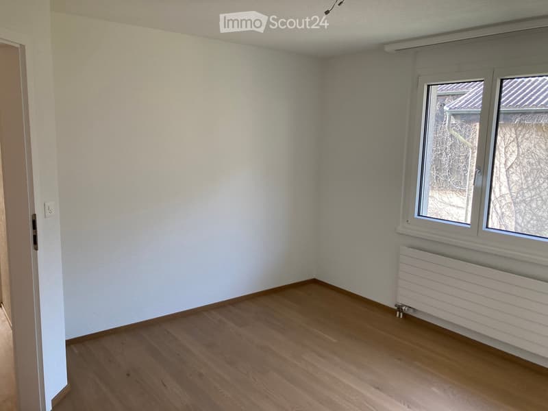 Empty room with wooden flooring, white walls, a window with a view of a roof, and a radiator below the window.