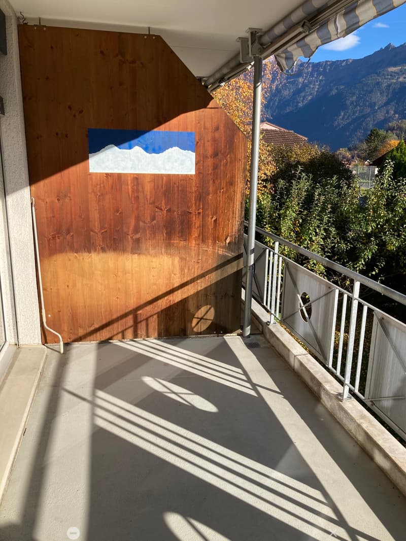 Balcony with concrete floor, metal railing, and a wooden door leading to the interior. The view shows mountains and greenery.