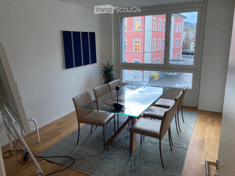 Dining room with a glass table, six chairs, a plant, and a painting on the wall. Large windows offer a view of the street.