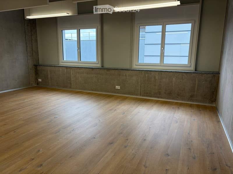 Empty room with wooden flooring, concrete walls, and large windows with blinds.