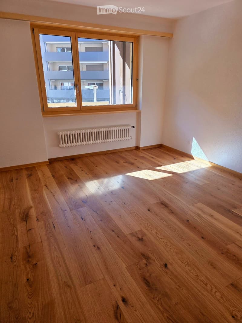 Empty room with wooden flooring, large window with wooden frame, radiator, and natural light coming in.