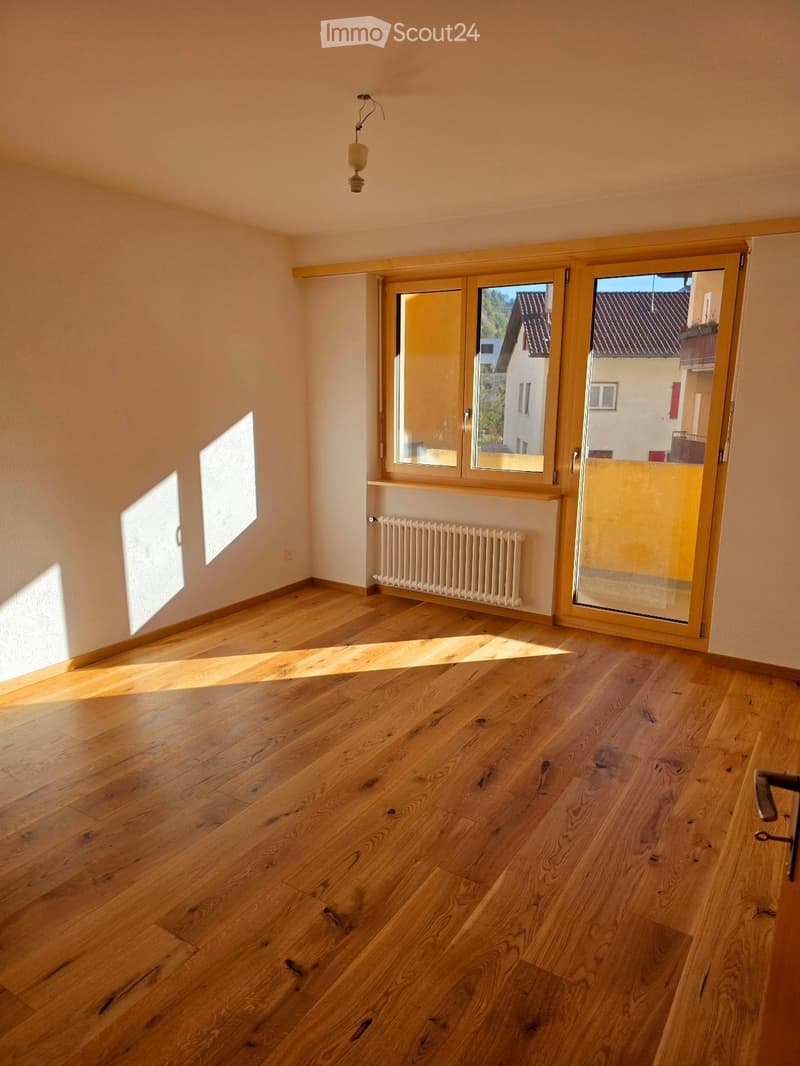 Empty room with wooden flooring, white walls, large windows, and a radiator. Sunlight streams in from the windows, casting shadows on the floor.