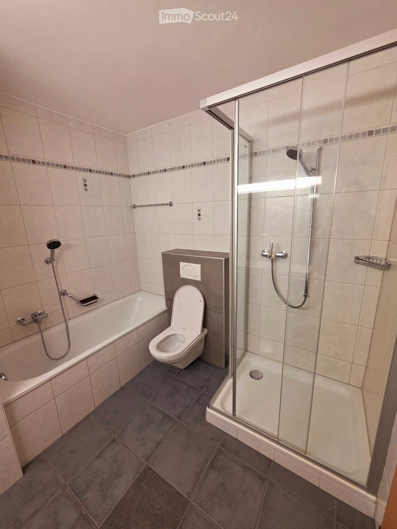 Modern bathroom with a bathtub, toilet, and walk-in shower with glass enclosure. Light-colored tiles and dark grey floor tiles.