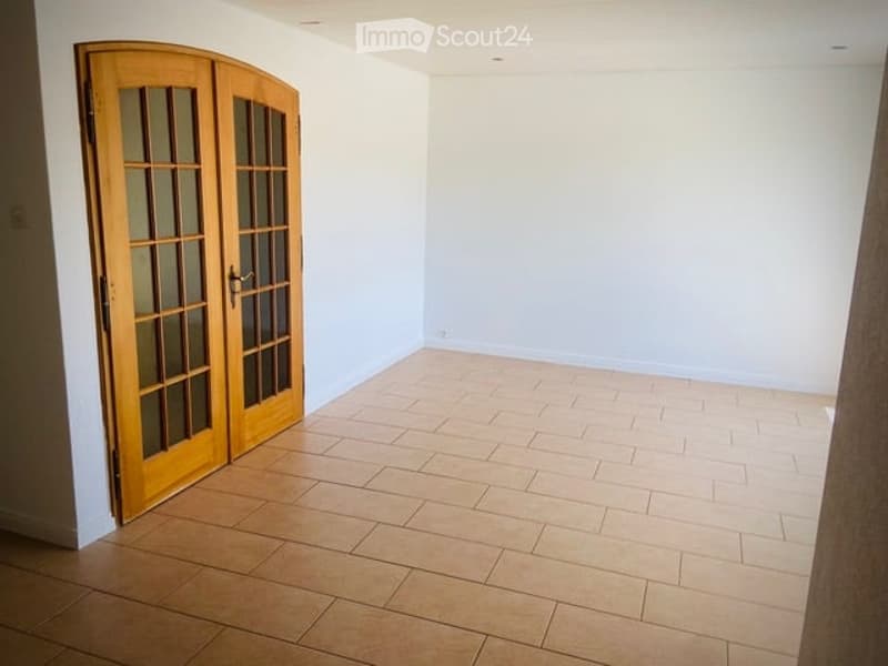 Empty room with light-colored tiled flooring and white walls, featuring a wooden double door with glass panels.