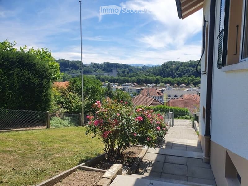 Garden area with a tiled pathway leading to a house, surrounded by greenery and flowers, with a scenic city view in the background.