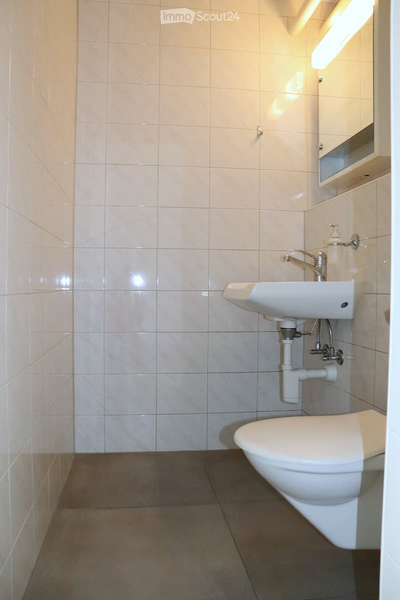 Modern bathroom with beige tiled walls and floor, featuring a white toilet, sink with chrome faucet, and a wall-mounted mirror with built-in shelf.