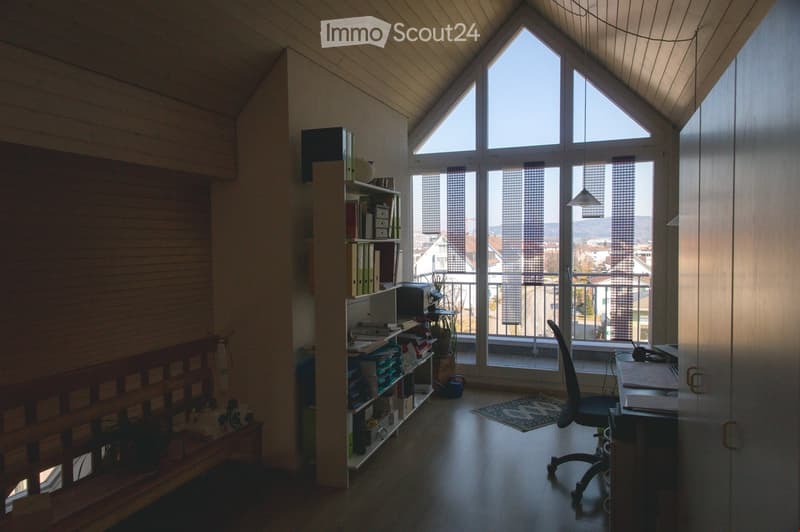 Bright room with wooden walls and ceiling, large windows with blinds, a desk with a chair, a bookshelf filled with books and office supplies, and a small plant on the floor.
