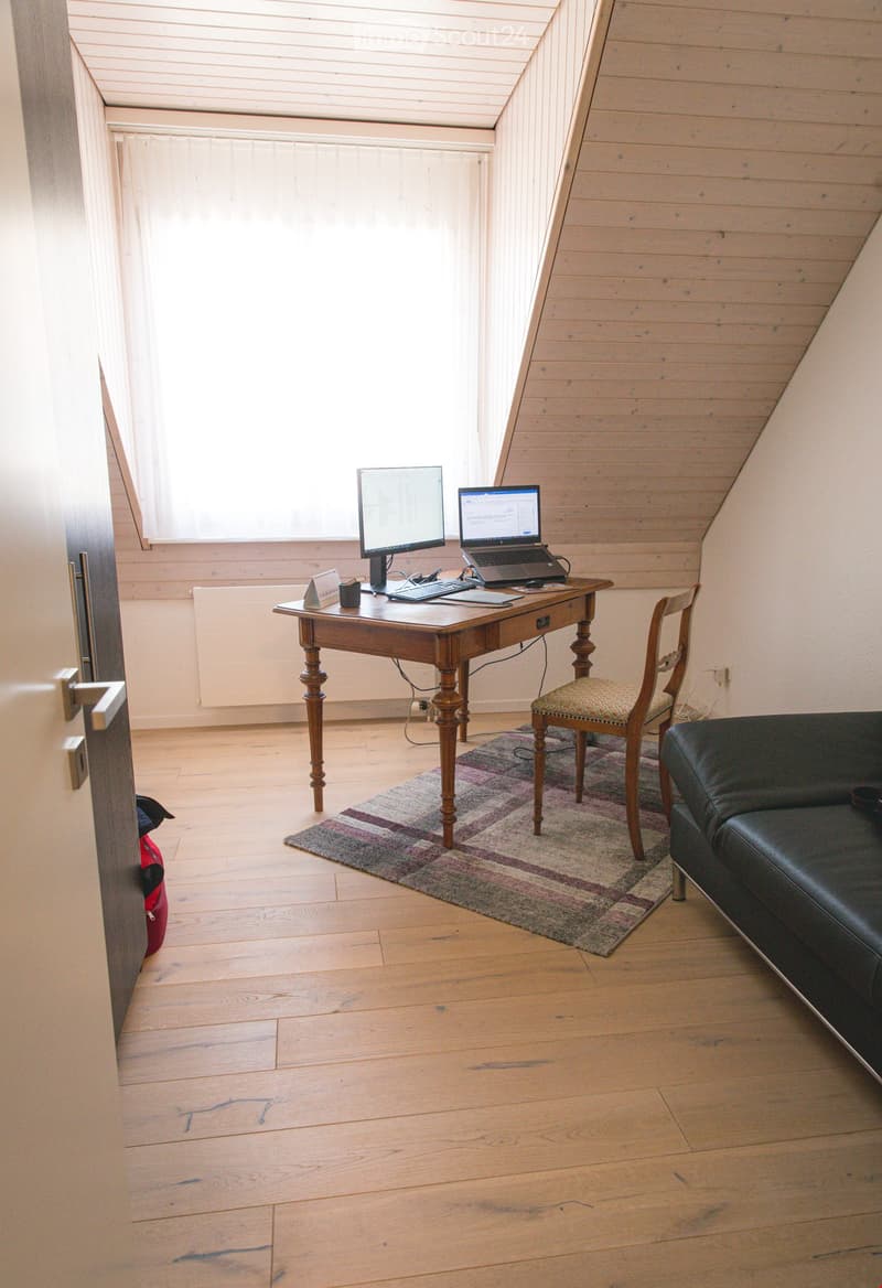 A cozy attic room with wooden floors, slanted wooden ceilings, and large windows. It features a wooden desk with a laptop, monitor, keyboard, and mouse, a patterned rug, a black leather sofa, and a wooden chair with a cushioned seat.