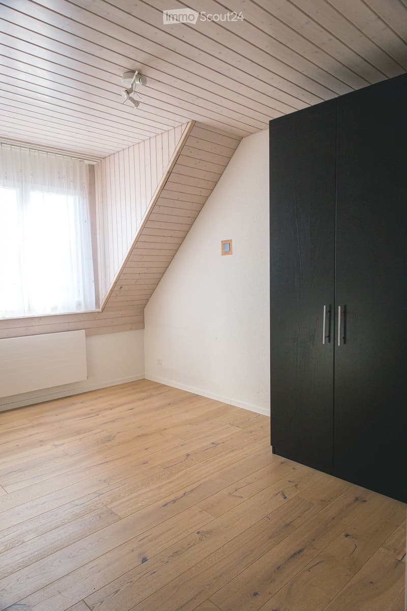 Empty attic room with wooden floors, white walls, and a slanted wooden ceiling. Features a large window with white curtains and a black wardrobe with silver handles.