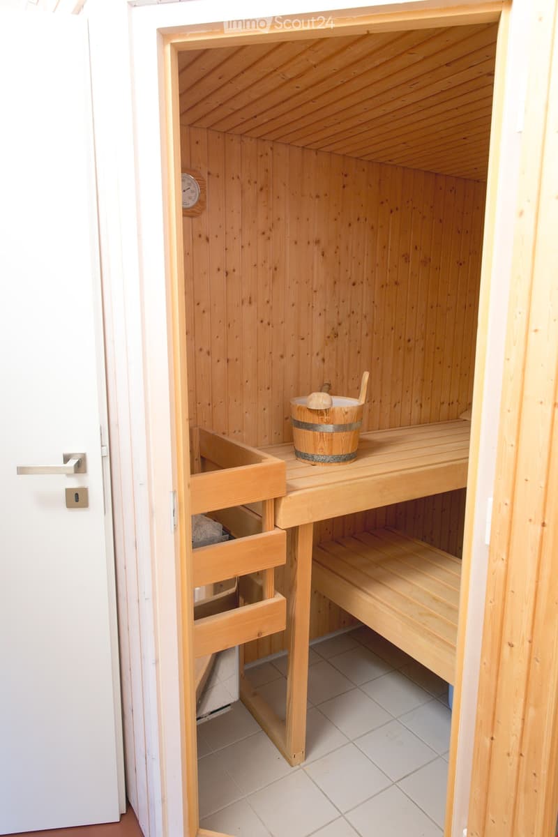 A small wooden sauna room with a wooden bench, a wooden bucket, and a wooden drawer. The room has light wooden walls and a tiled floor.